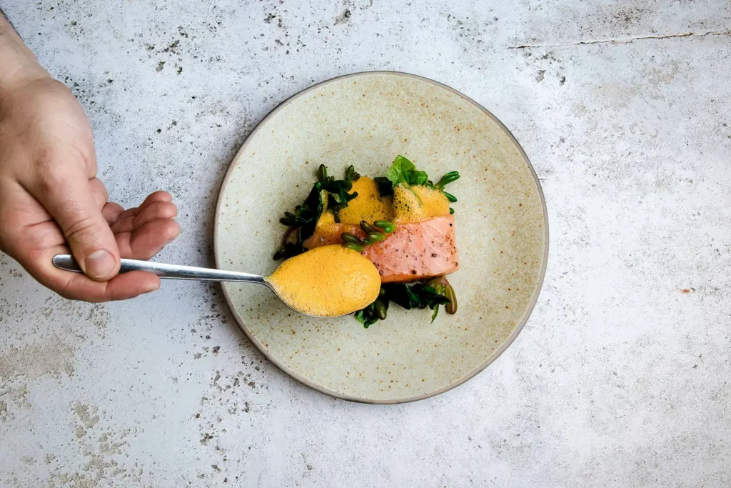 cooking_image_29 Der Privatkoch hält eine Gabel über einen Teller mit Essen und ist bereit, und garniert das Essen mit einer schaumigen Soße.